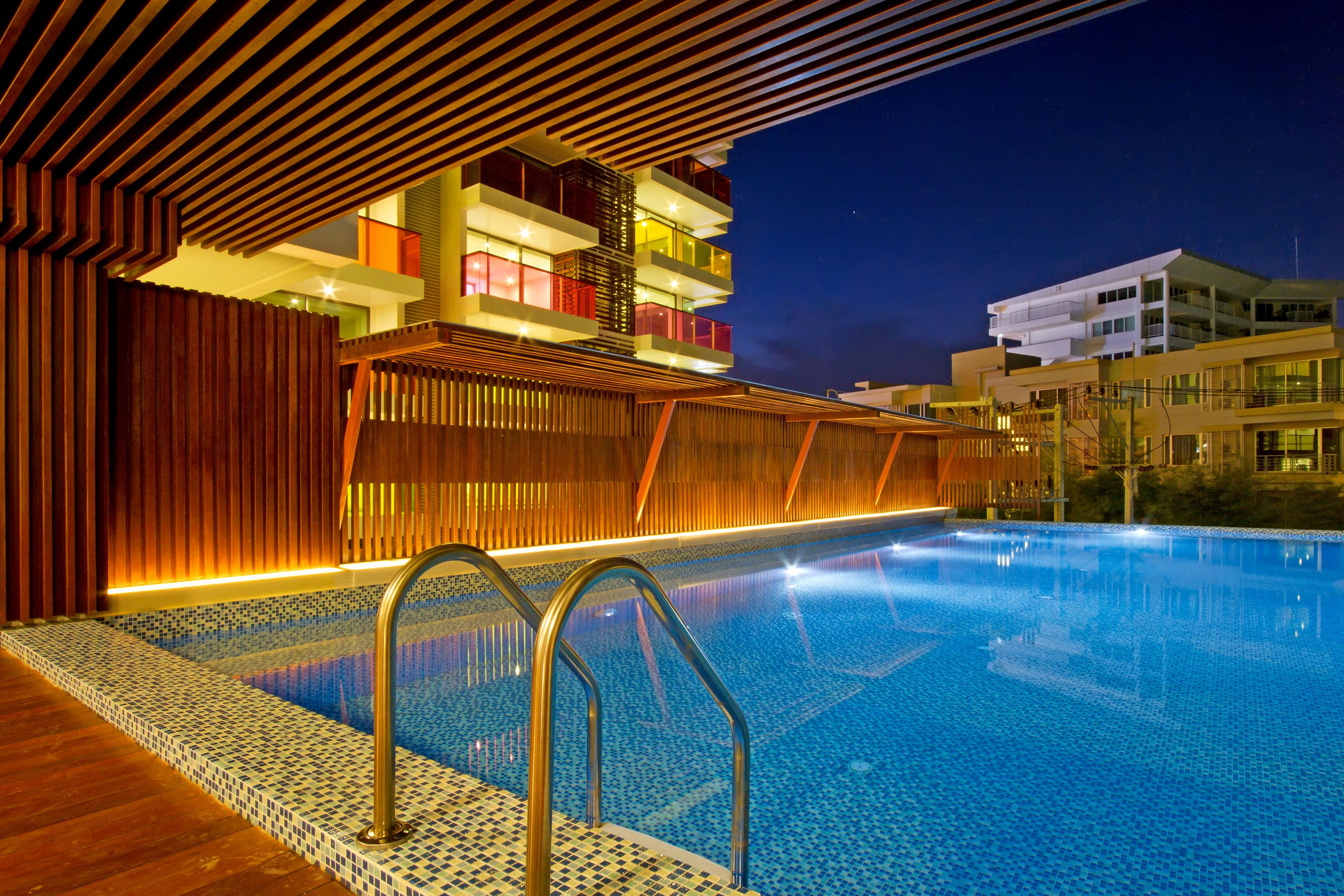 A swimming pool with a wooden deck and metal railings, surrounded by modern buildings with balconies and illuminated windows.