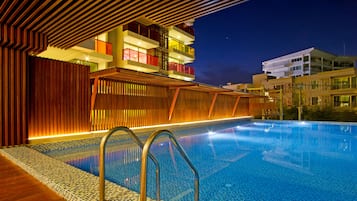 A swimming pool with a wooden deck and metal railings, surrounded by modern buildings with balconies and illuminated windows.
