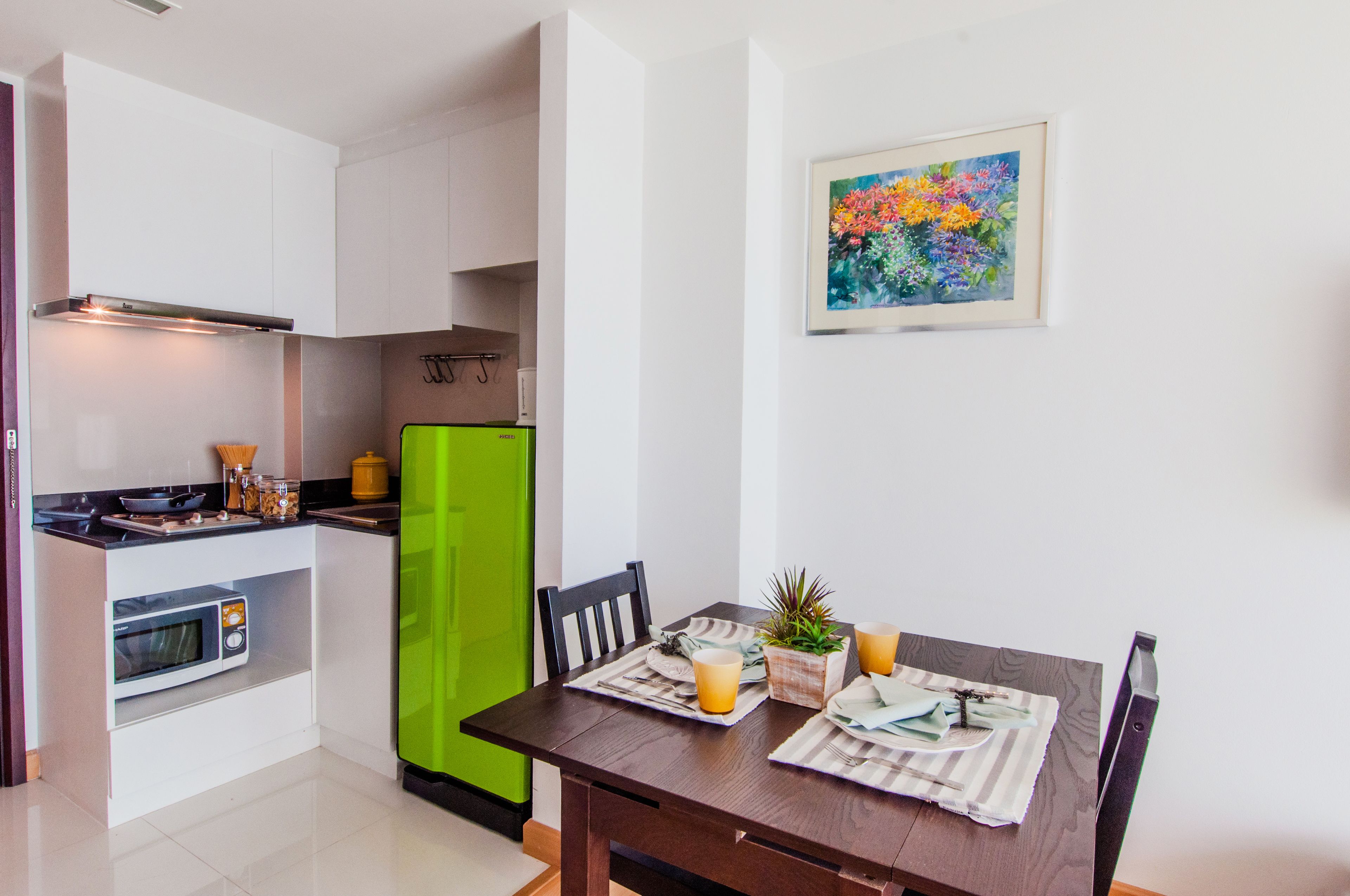 Una cocina compacta con armarios blancos, un microondas y un refrigerador verde. Una mesa de comedor con un mantel individual y una planta en maceta, dispuesta con un vaso de jugo de naranja.