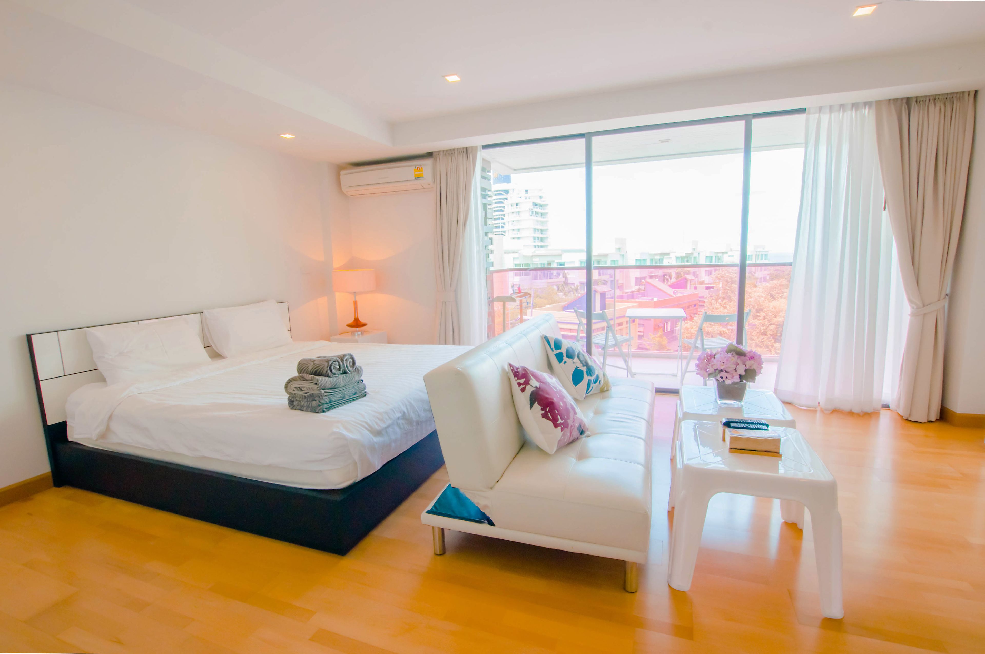 A modern hotel room with a large bed, a sofa, a small table with a vase, and a view of the city through large windows.