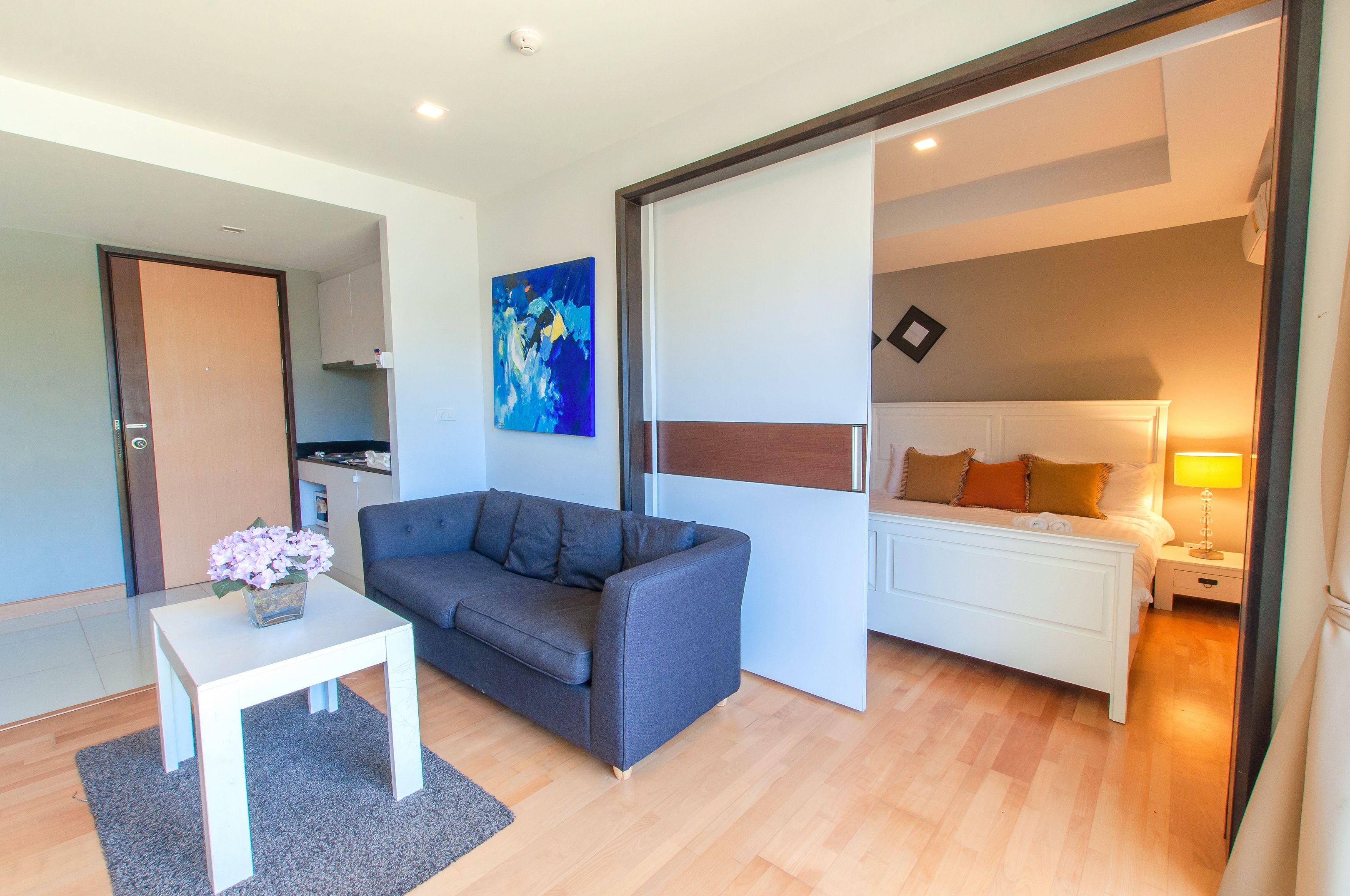 Ein modernes Wohnzimmer mit einem grauen Sofa, einem kleinen weißen Tisch mit Blumenvase und einem Bett mit Nachtschränkchen.