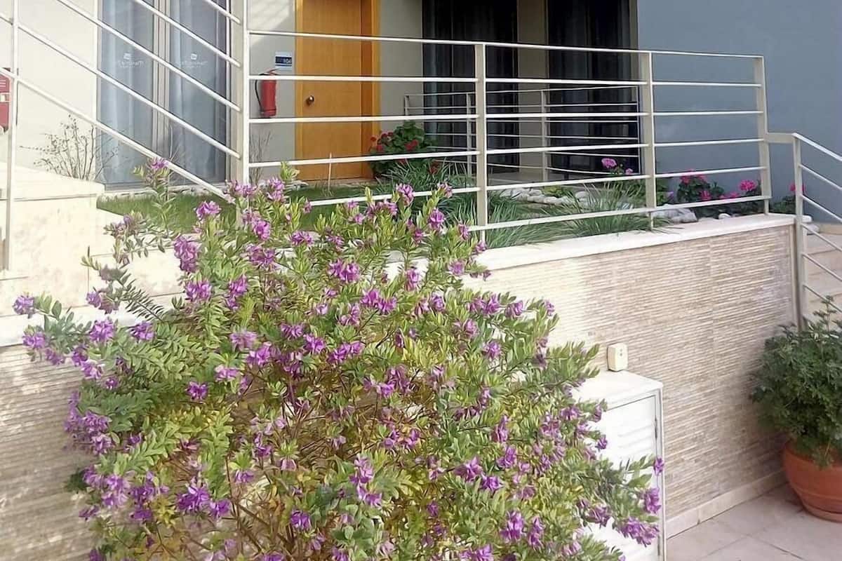 A modern two-story residential building with a balcony, a wooden door, and a small garden area with purple flowers.