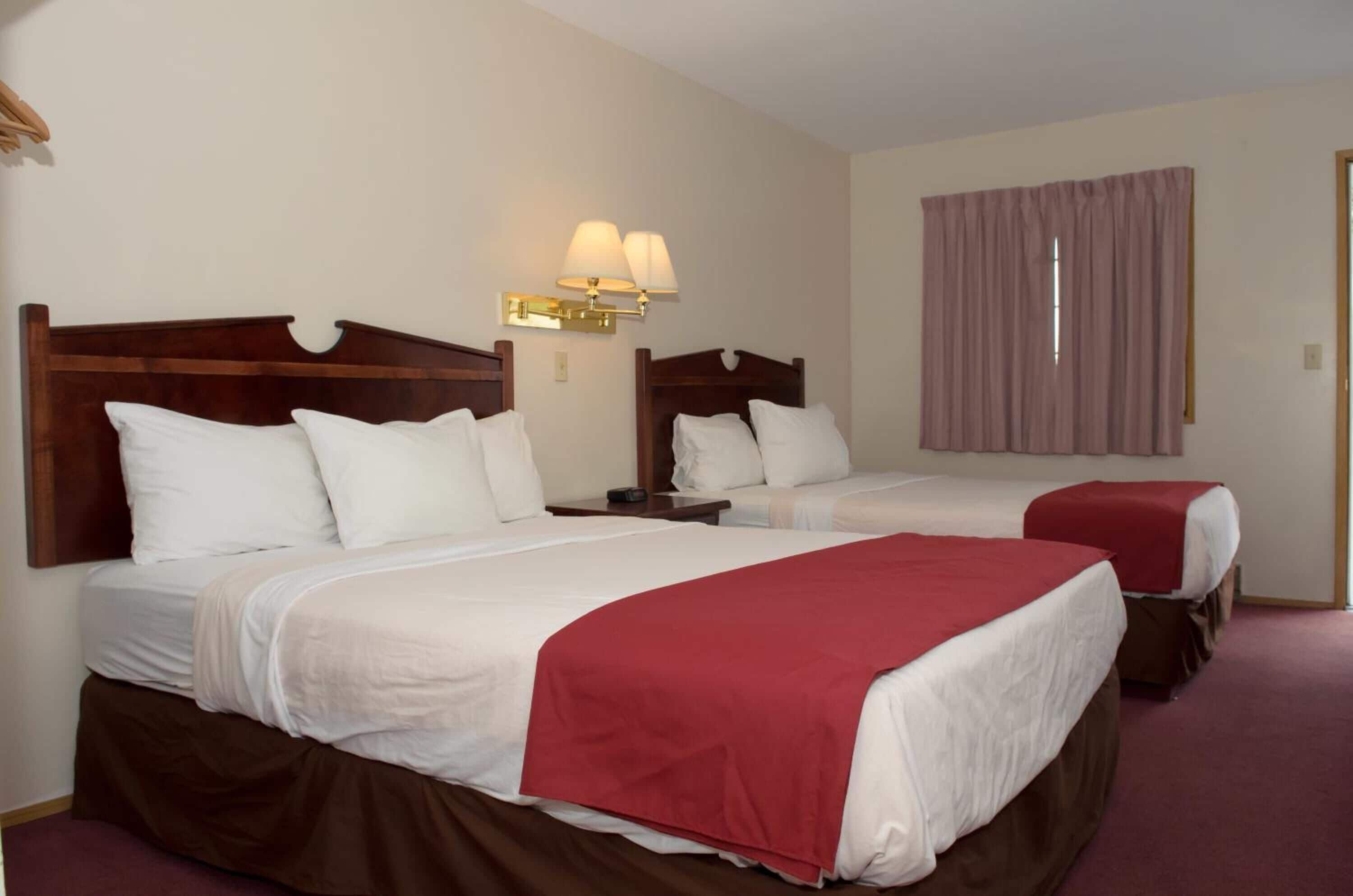 A hotel room with two beds, each with white bedding and a red blanket. There is a wall-mounted lamp above the beds and a window with curtains.