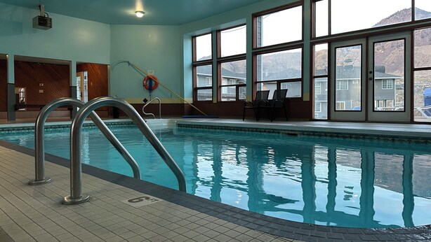 An indoor swimming pool with clear blue water, a metal ladder, and large windows offering a view of the surrounding landscape. - Kings Motor Inn (Kamloops)