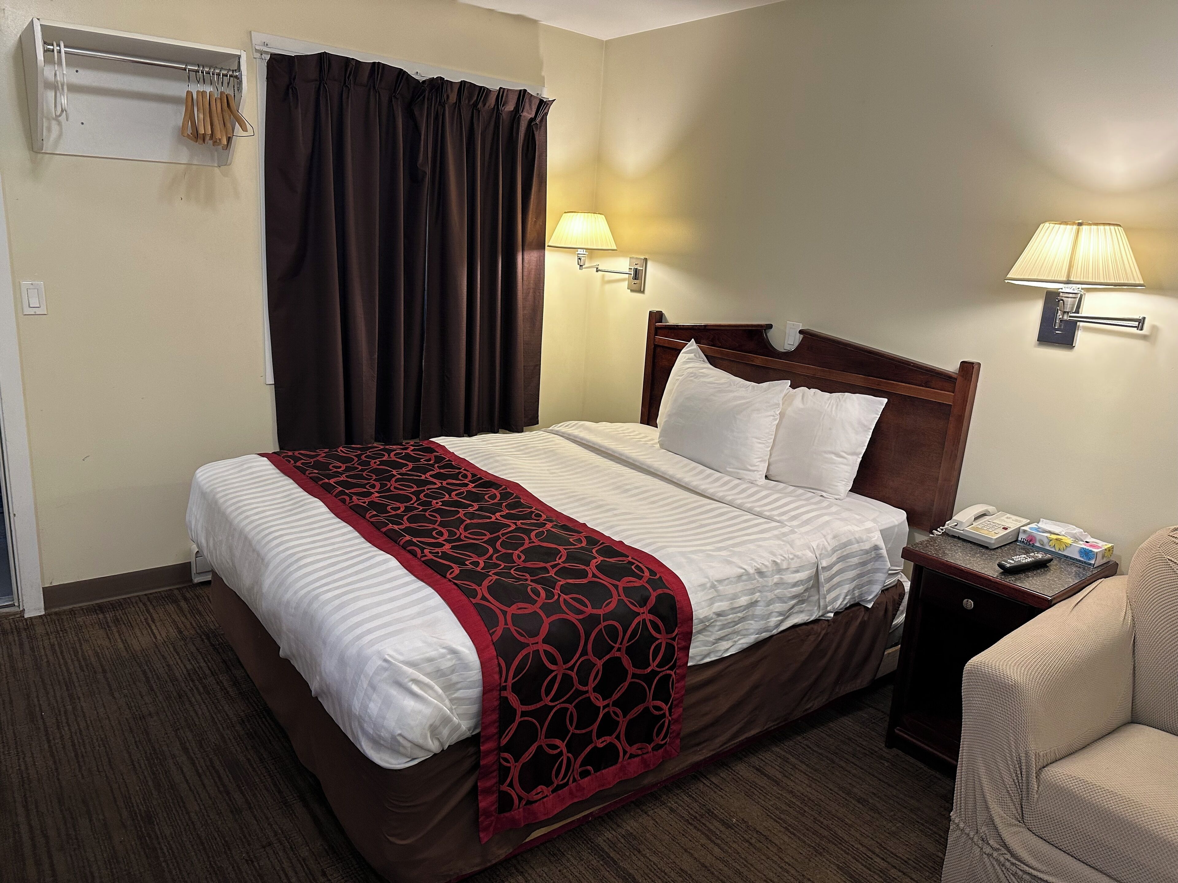 A hotel room with a bed, a bedside table with a lamp, a chair, and a coat rack with hangers.