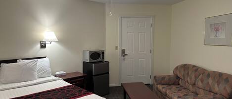 A hotel room with a bed, a desk, a microwave, a refrigerator, a sofa, a ceiling fan, and a door.
