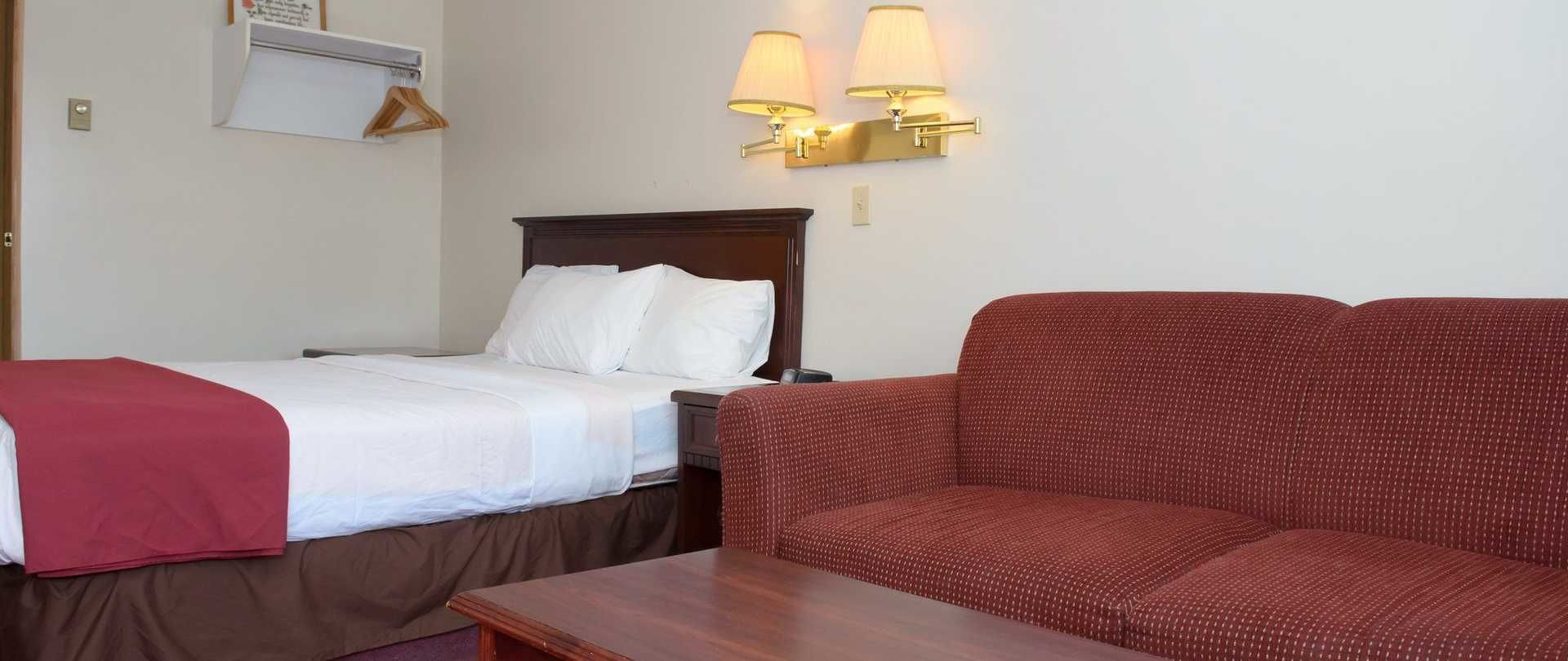 A hotel room with a bed, a red sofa, and a small table. There are two wall-mounted lamps and a coat rack with hangers.