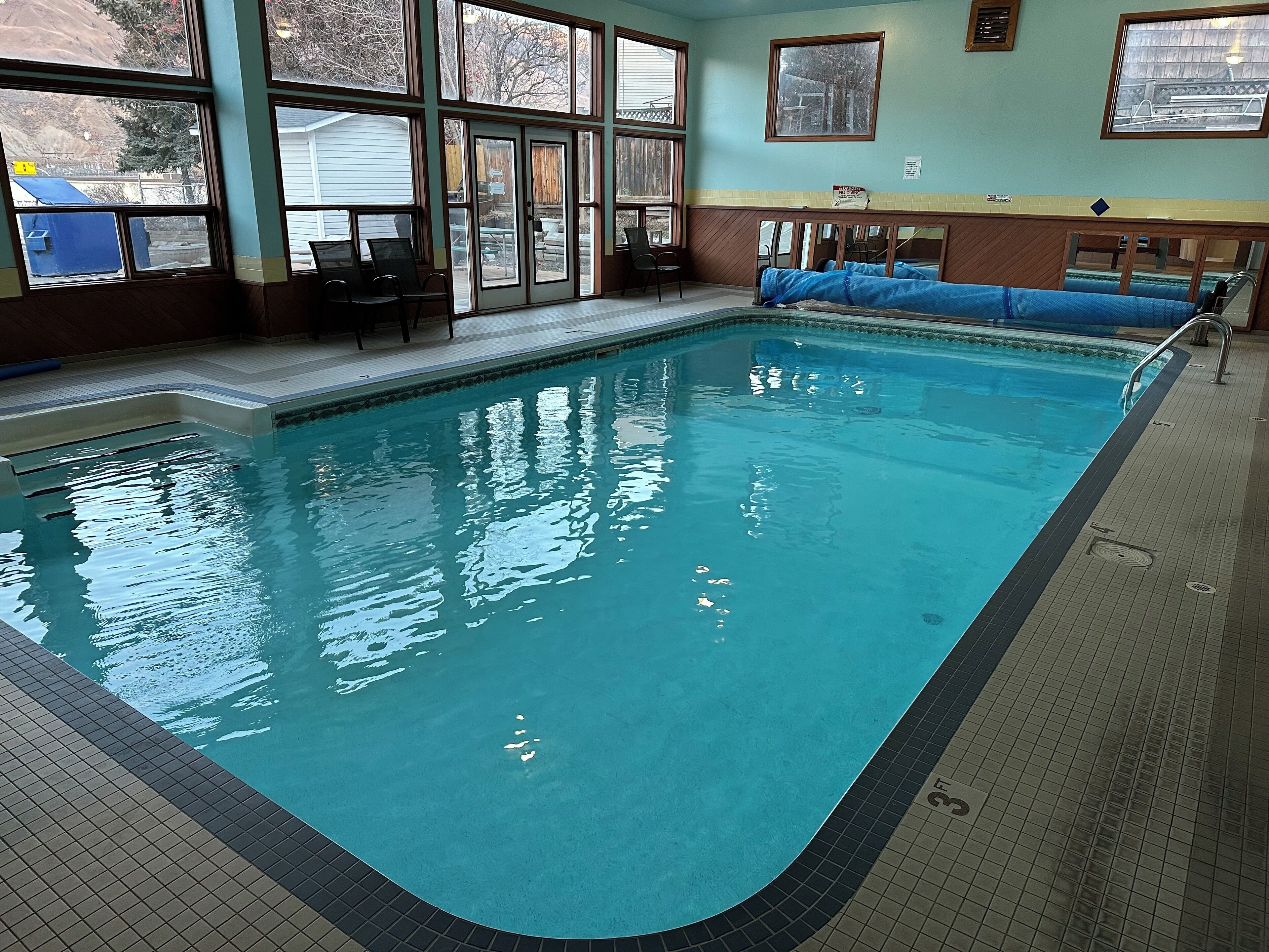 Ein Schwimmbecken mit klarem, blauem Wasser, umgeben von einer gefliesten Terrasse und großen Fenstern.