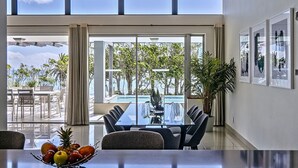A modern dining area with a glass table and chairs, a fruit bowl, and a view of an outdoor pool and trees. - Blue Views Penthouse Beachfront Apartments (Cape Town)