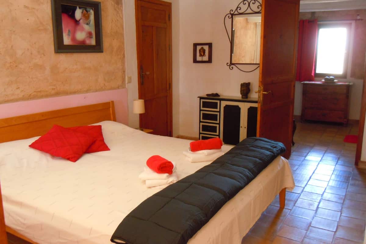 A neatly made bed with red pillows and a black blanket, a wooden dresser, a mirror, and a red door leading to another room.