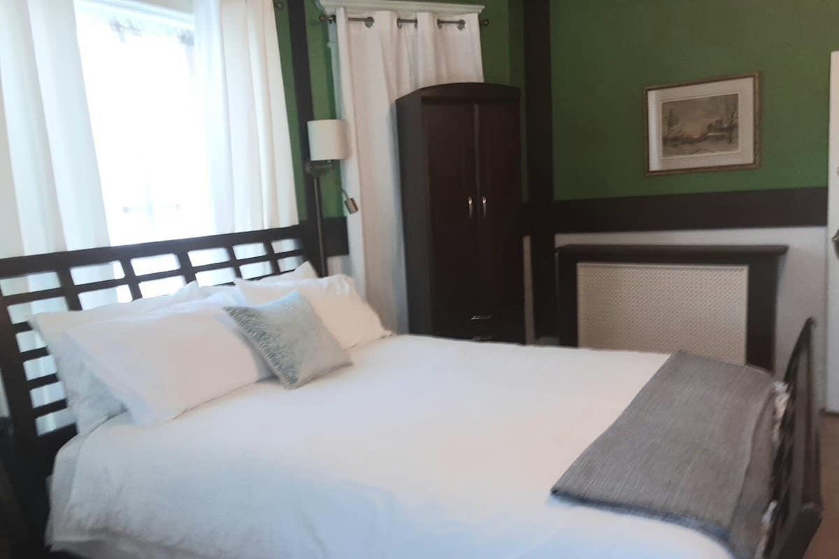Un lit bien fait, avec du linge de lit blanc et un plaid gris, une armoire en bois foncé et un tableau encadré accroché au mur.