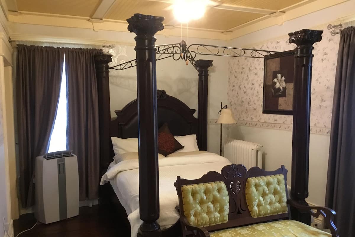 Un lit à baldaquin avec une structure en bois foncé, un couvre-lit blanc et un seul oreiller rouge. On aperçoit une chaise en bois avec un siège et un dossier rembourrés de tissu jaune. Un radiateur est visible sur le mur, et une fenêtre avec des rideaux se trouve sur le côté gauche.