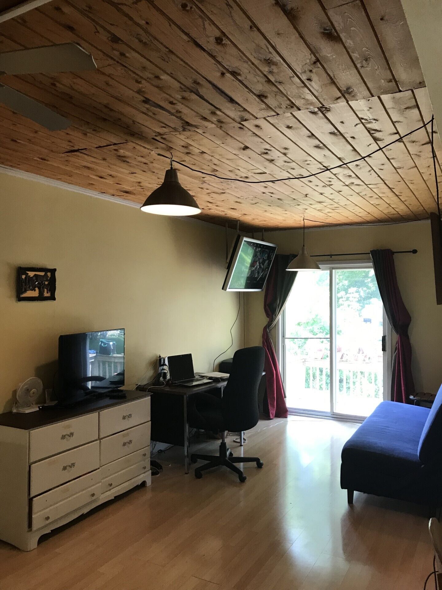 Een kamer met een houten plafond, een bureau met een computer, een televisie, een bed en een raam met gordijnen.