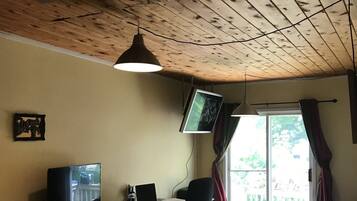 Een kamer met een houten plafond, een bureau met een computer, een televisie, een bed en een raam met gordijnen.