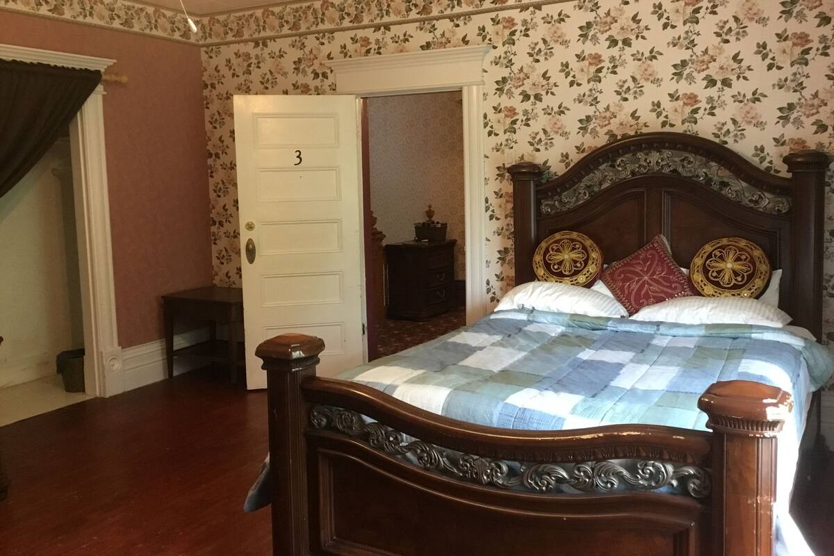 Une chambre à coucher avec un lit en bois, un papier peint à motifs floraux, une porte portant l’inscription « 3 », et une petite table de chevet.