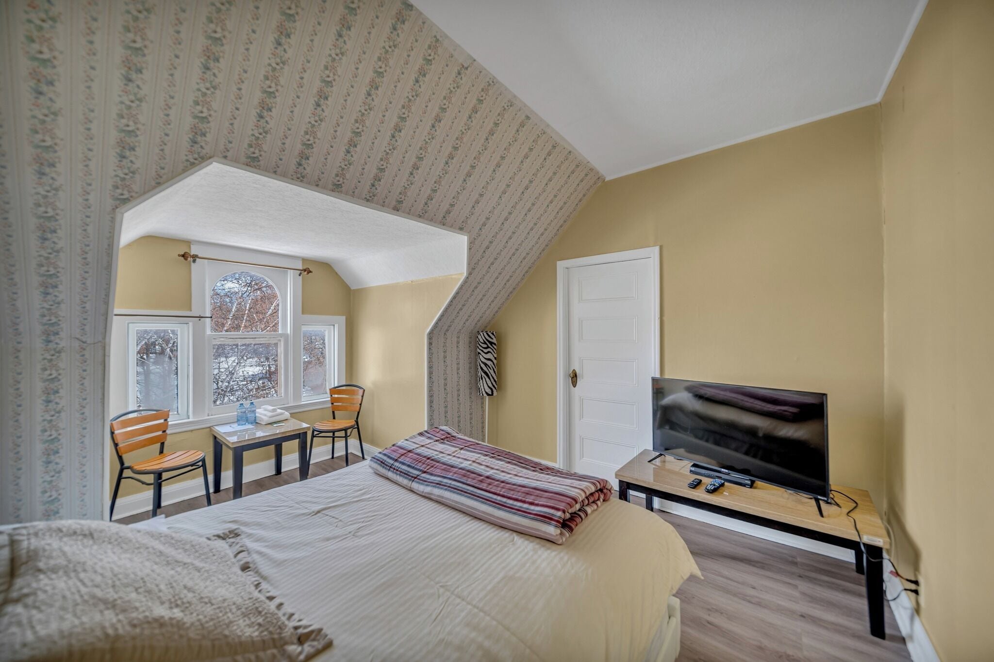 A bedroom with a bed, a television, a desk with a chair, and a window with a view of trees.