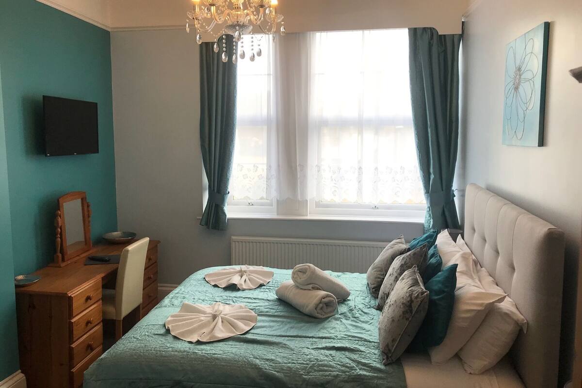 Un dormitorio con cama, cómoda, televisor, candelabro y ventana con cortinas.