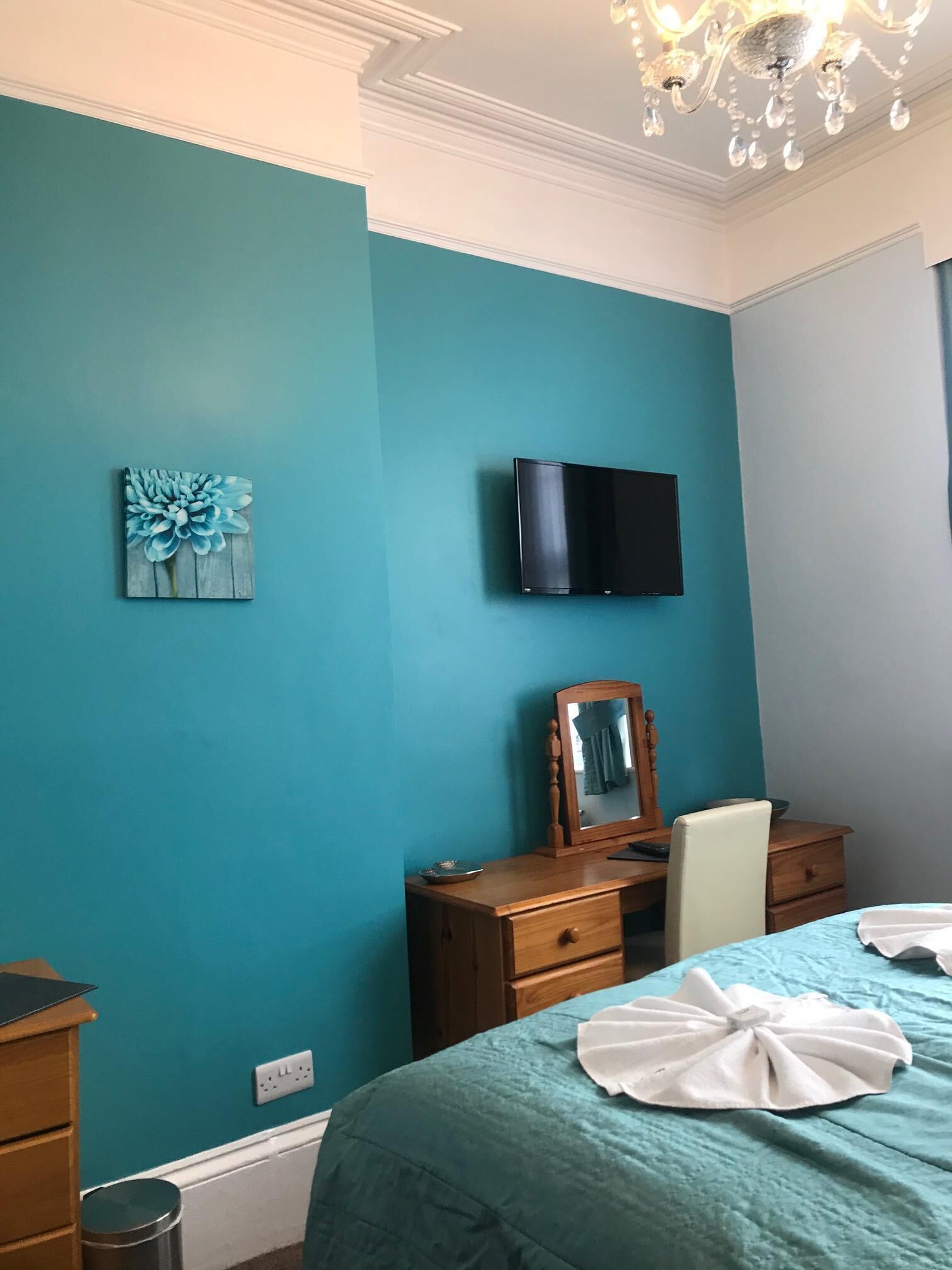 Un dormitorio con paredes color turquesa, una cama con toallas blancas, un aparador de madera, un espejo y un televisor de pantalla plana montado en la pared.