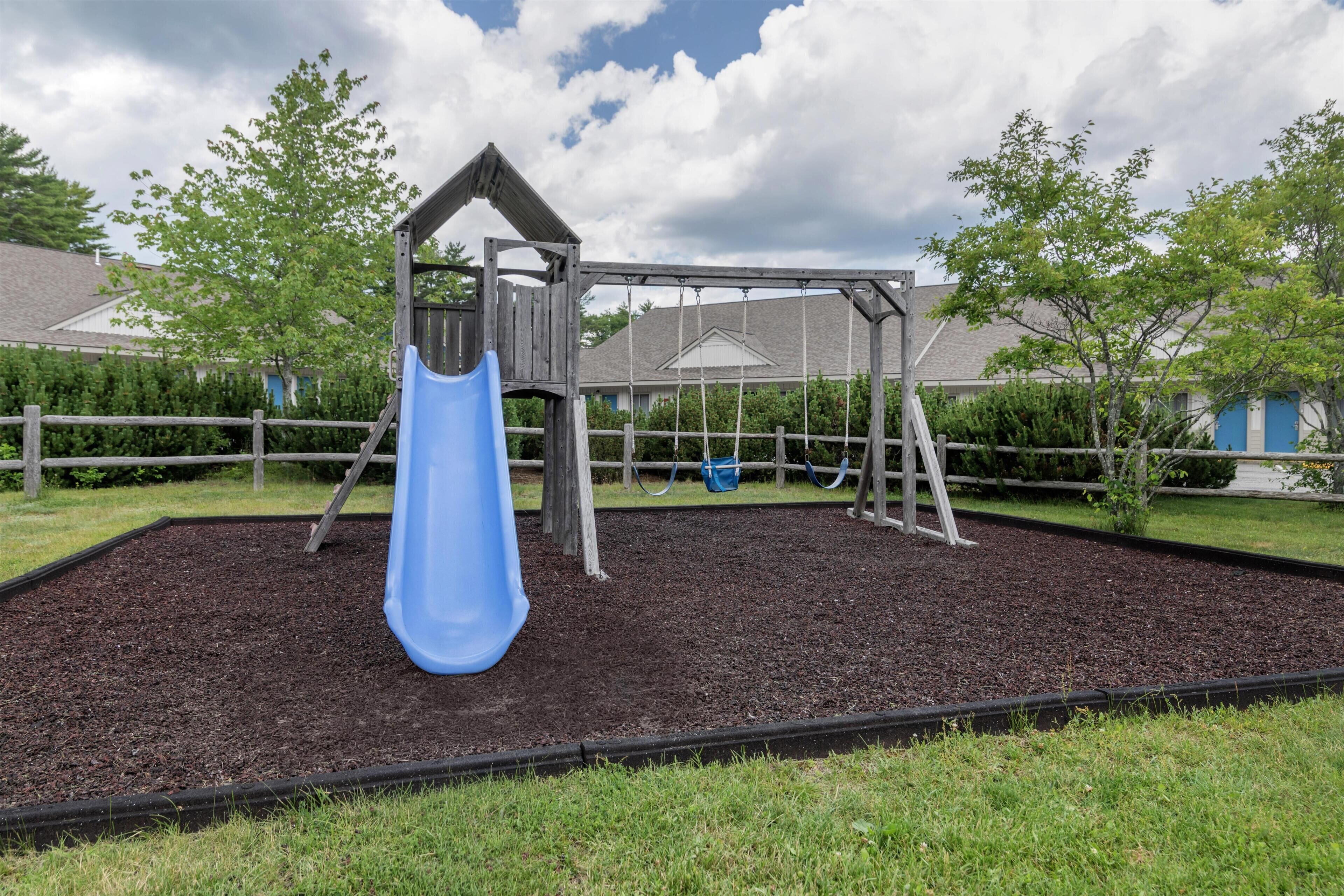 A playground with a slide and swings, surrounded by a fence and mulch, with houses in the background.