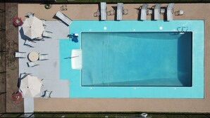 Vue aérienne d’une piscine entourée de chaises longues et de parasols, elle-même bordée d’une terrasse en briques et d’une clôture en métal.
