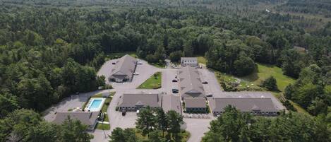An aerial view of a large, multi-building complex surrounded by dense forest, with a central parking area and a swimming pool.