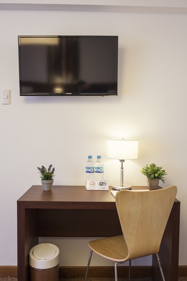 A hotel room with a flat-screen TV mounted on the wall, a desk with a lamp, two potted plants, a trash bin, and a water bottle.