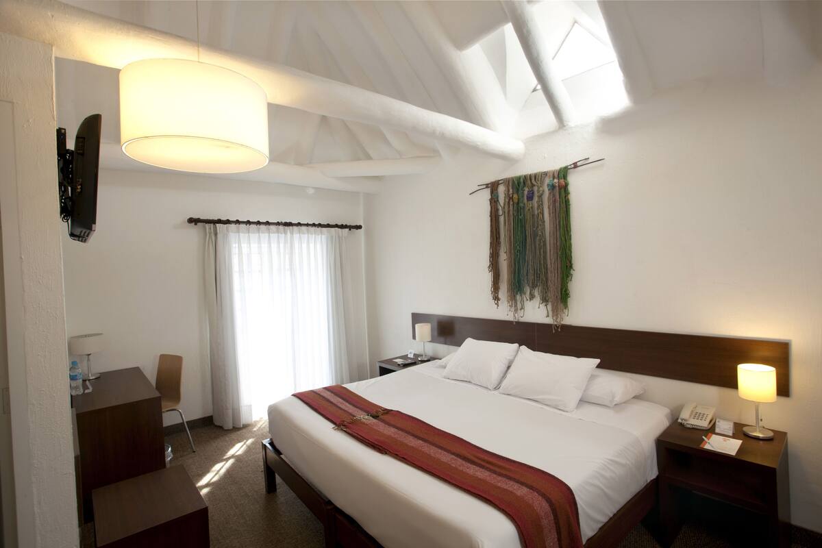 Una habitación de hotel con una cama grande, un escritorio, una silla, una mesita de noche con lámpara y un teléfono instalado en la pared.