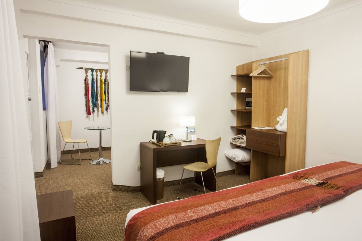 Habitación de hotel con cama, escritorio, silla, televisor, armario y una mesita de noche con lámpara.