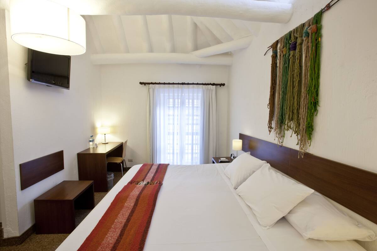 Habitación de hotel con una cama grande, un escritorio, una silla, un televisor montado en la pared y una ventana con cortinas.