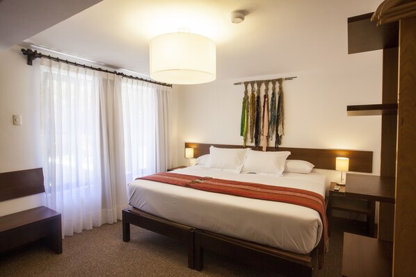 Habitación de hotel con cama, lámparas de noche, un banco y un ventanal con cortinas translúcidas.