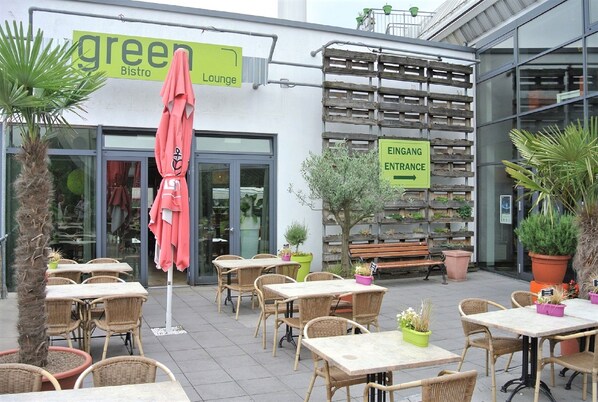 An outdoor cafe area with wicker tables and chairs, potted plants, and a sign indicating the entrance to a building.