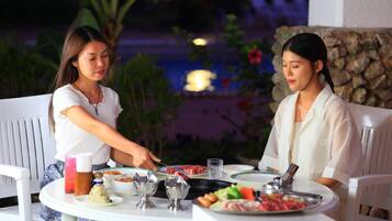 Two people dining at an outdoor table with a variety of dishes, including meat and vegetables, in a garden setting with a stone wall and plants in the background.