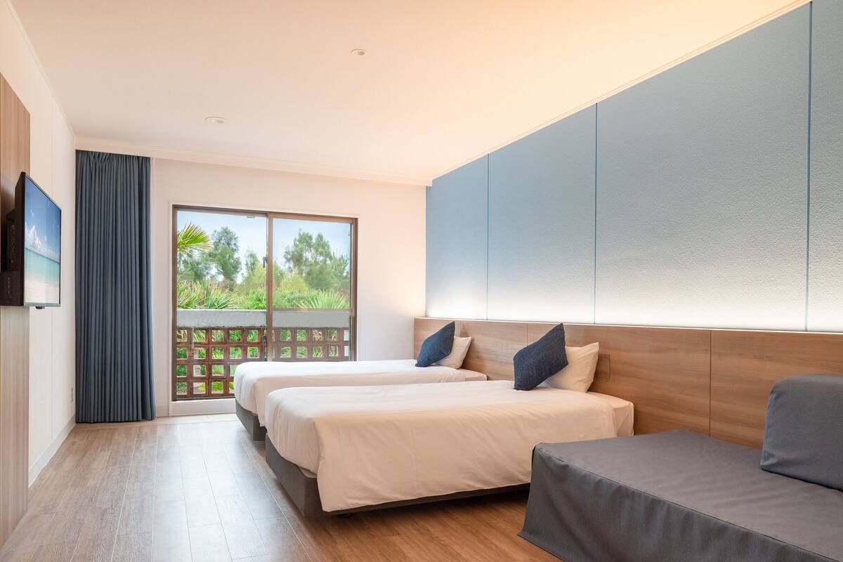 Une chambre d’hôtel moderne équipée de deux lits, d’un canapé, d’une télévision et d’un balcon offrant une vue sur la verdure.