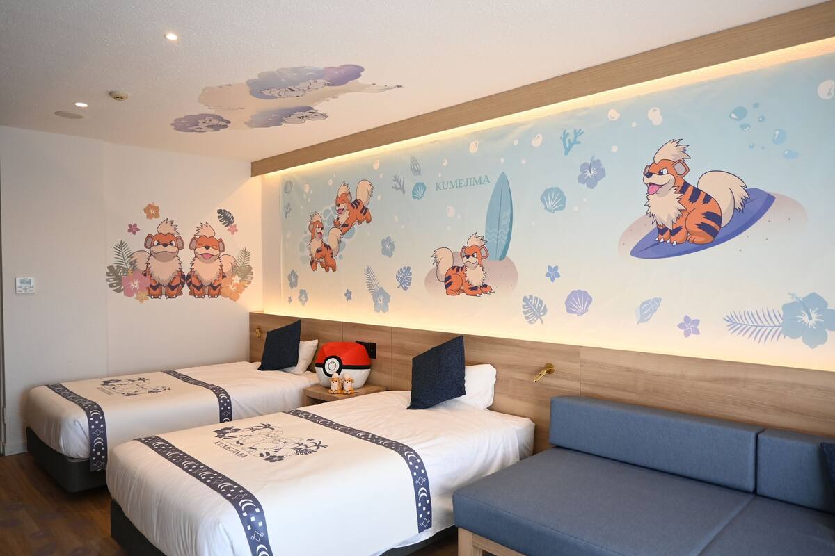Une chambre d’hôtel avec deux lits, un canapé et un mur décoré de personnages de dessins animés, ainsi que le nom « KUMEJIMA ».
