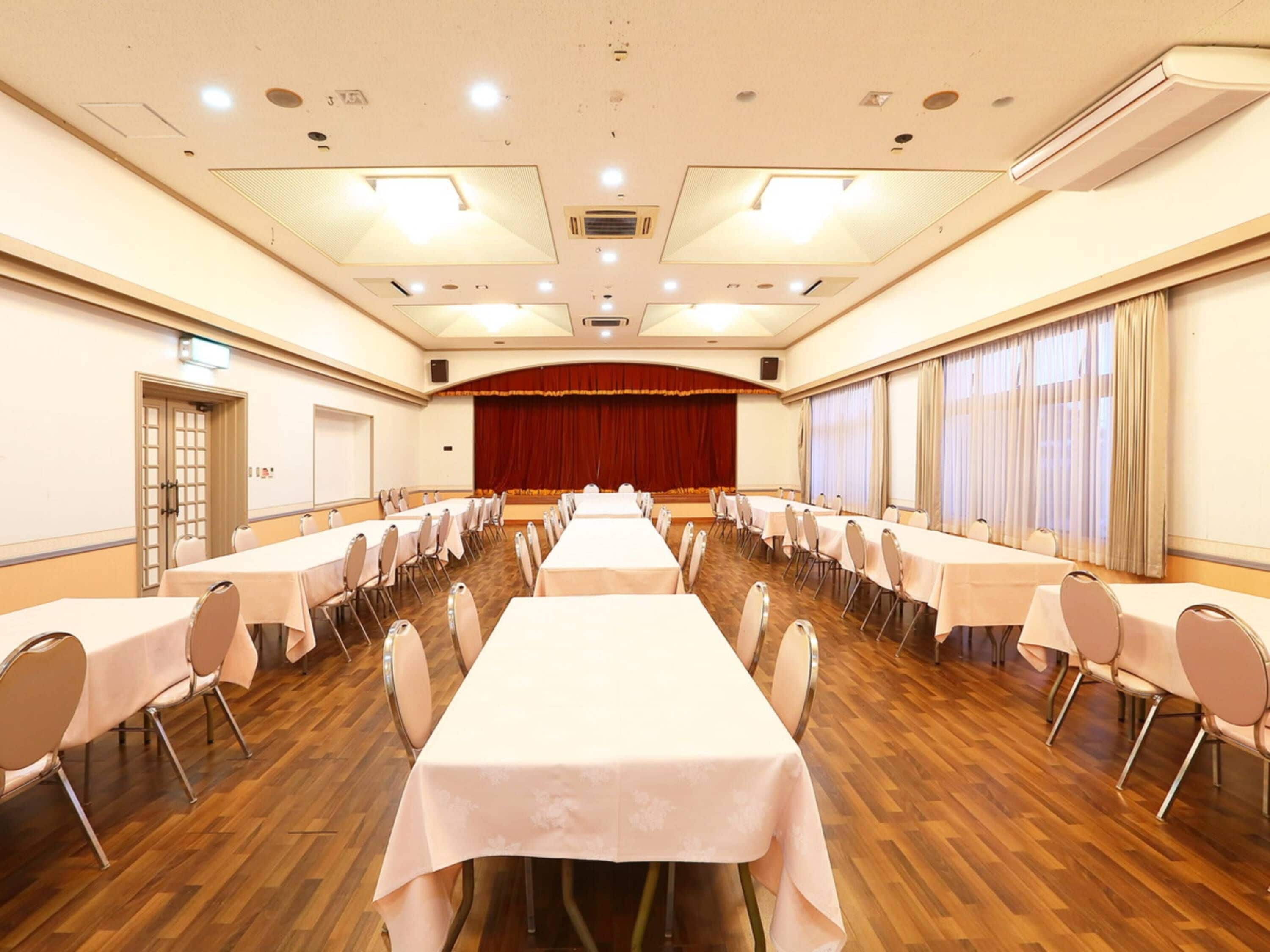 A conference room with multiple tables and chairs, wooden flooring, and a stage with curtains.