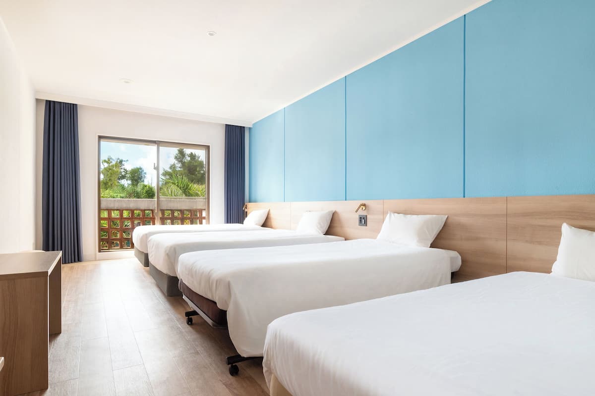 Une chambre d’hôtel avec trois lits, un bureau en bois et une vue sur la verdure par la fenêtre.