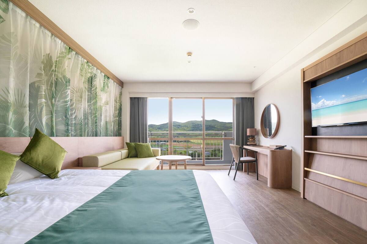 Une chambre d’hôtel dotée d’un grand lit, d’un canapé, d’un bureau avec une chaise, d’une télévision et d’un balcon offrant une vue sur le paysage.