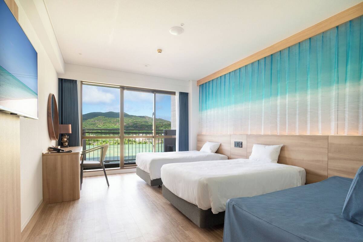 Une chambre d’hôtel avec deux lits, un bureau, une chaise, une télévision et un balcon offrant une vue sur les montagnes.