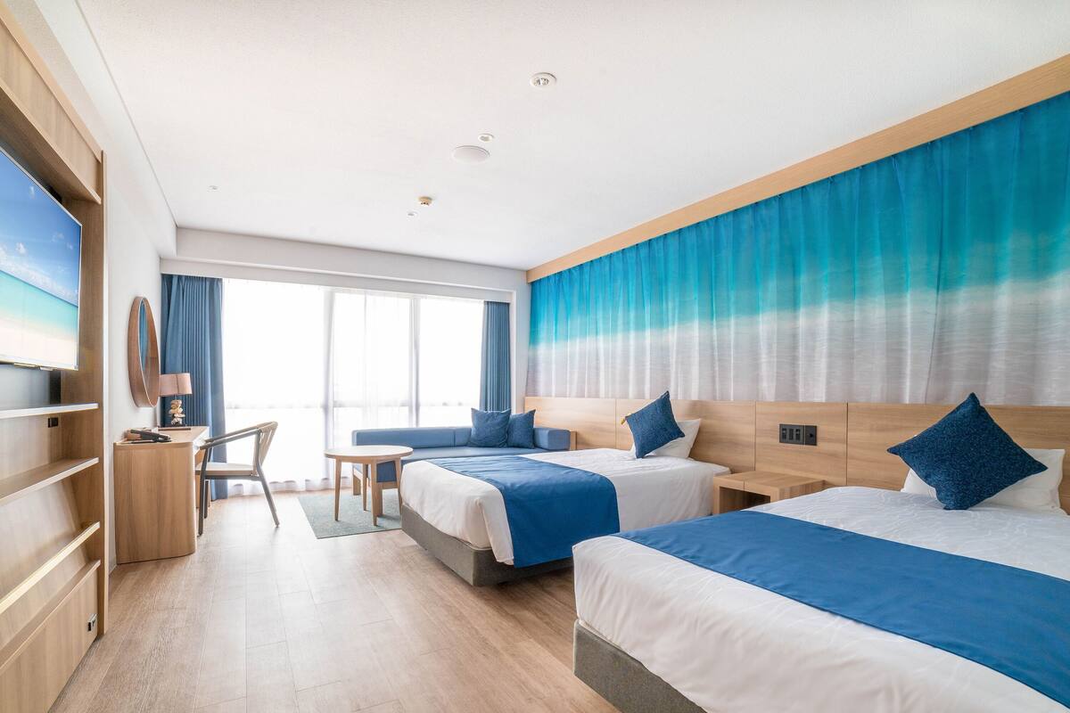 Une chambre d’hôtel avec deux lits, un bureau, une chaise et une grande fenêtre avec des rideaux.