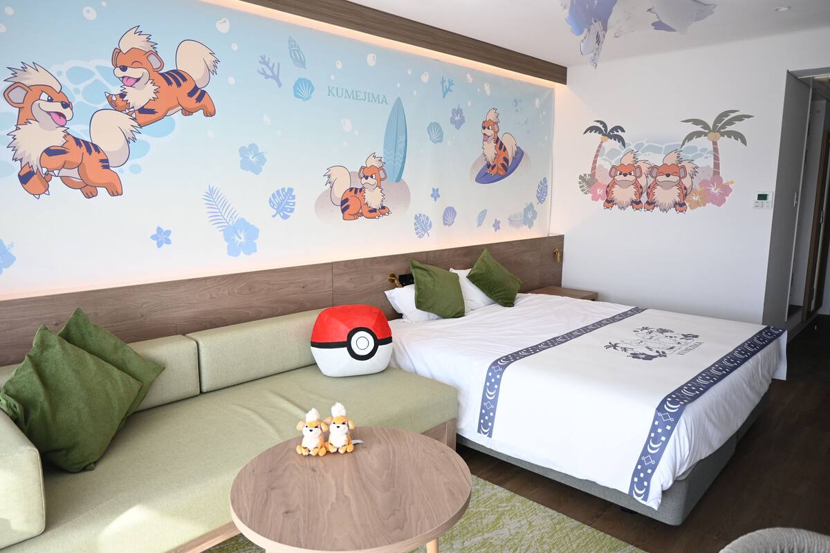 Une chambre d’hôtel avec un lit, un canapé vert et une table ronde en bois sur laquelle sont posés deux jouets en peluche.