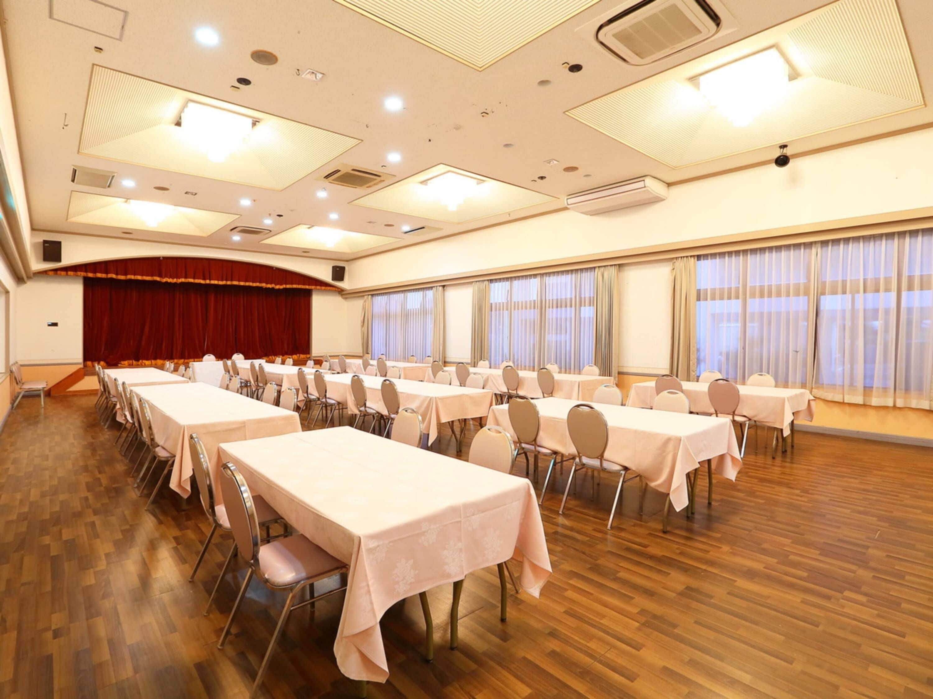 A conference room with multiple tables and chairs, a stage with curtains, and large windows with curtains.