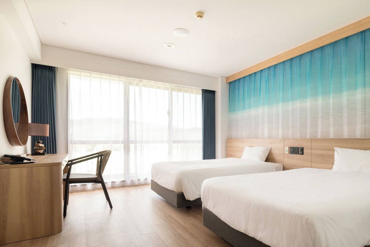 Une chambre d’hôtel avec deux lits, un bureau, une chaise et une grande fenêtre avec des rideaux.