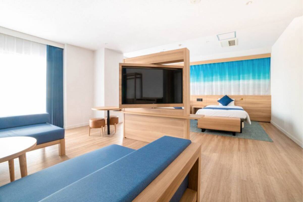Une chambre d’hôtel moderne dotée d’un grand lit, d’un téléviseur fixé sur une cloison en bois, d’un coin salon avec un banc et d’une grande fenêtre avec des rideaux.
