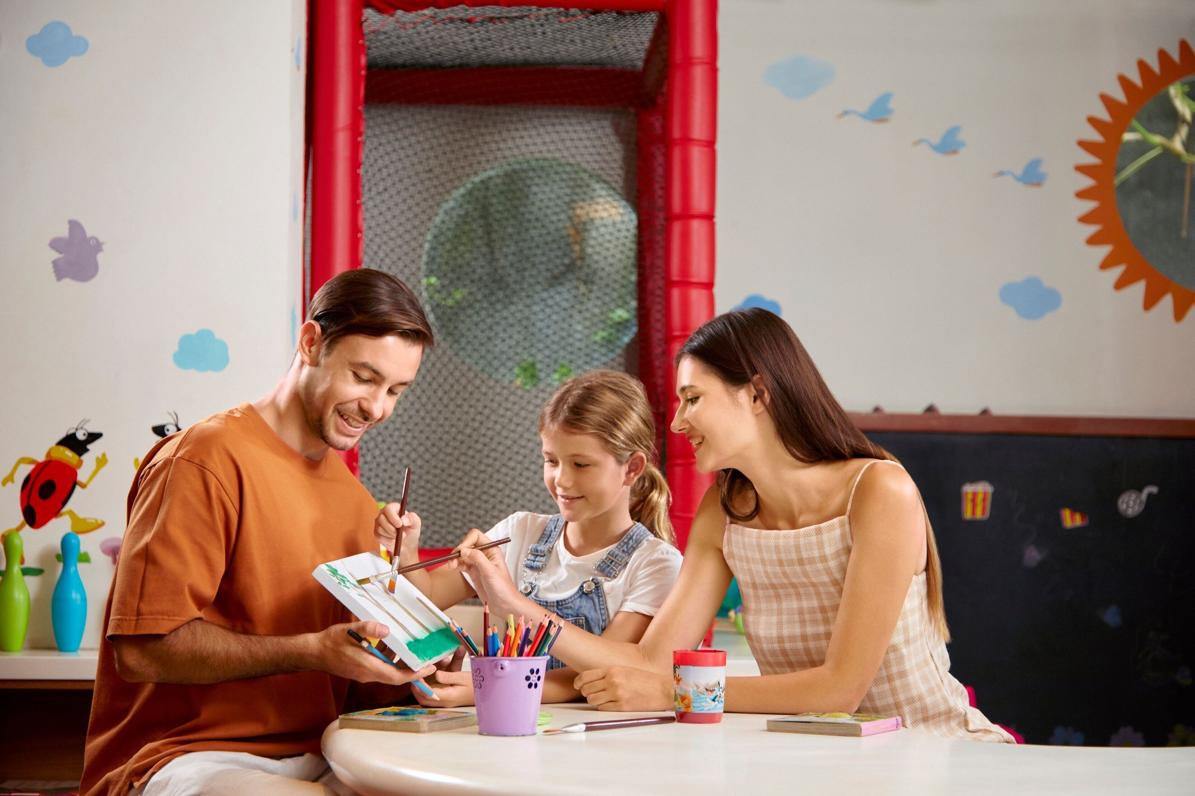 A family engaged in a painting activity at a table with art supplies, in a room with educational decorations and a play area visible in the background.
