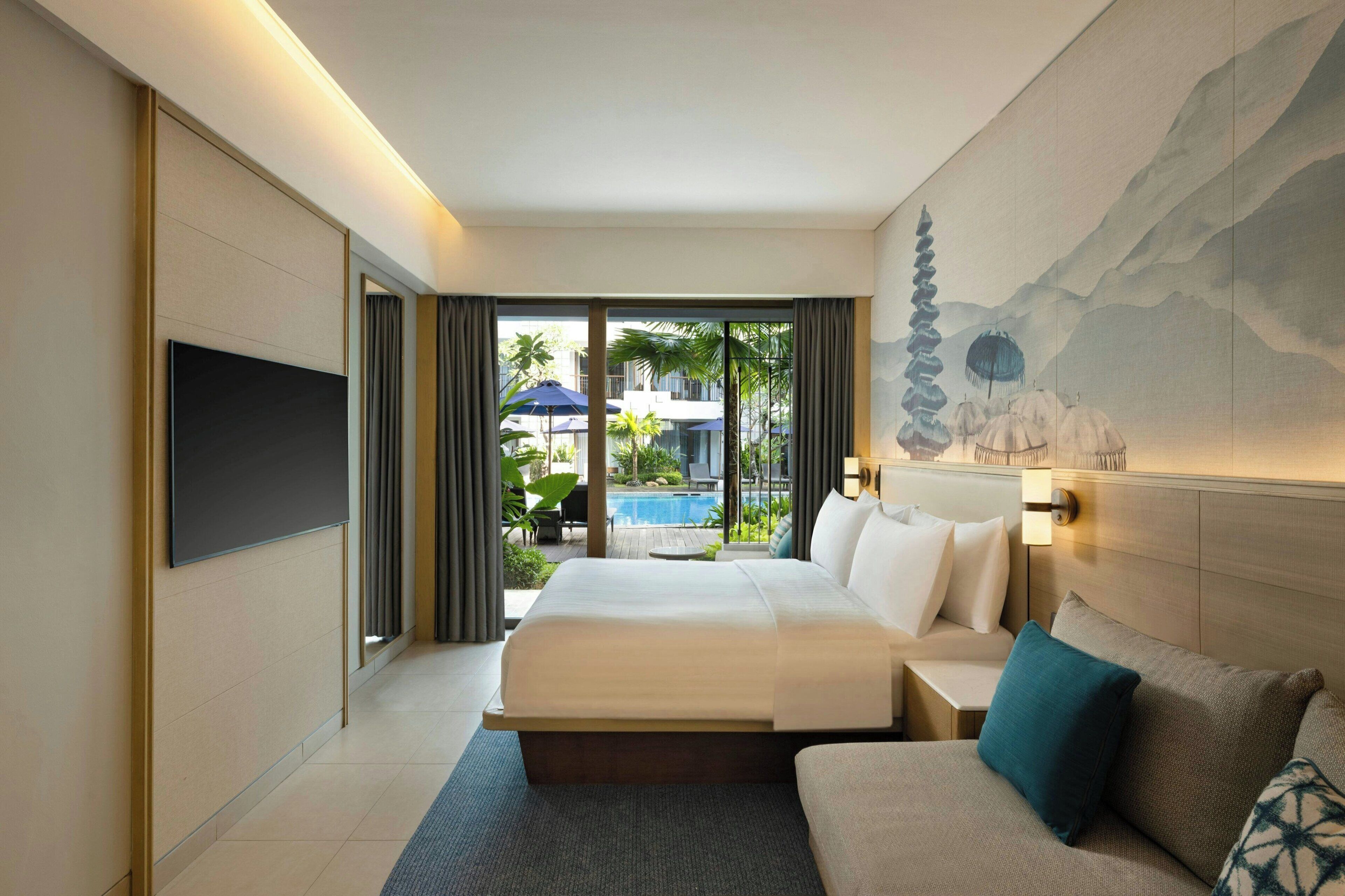 A modern hotel room with a large bed, a sofa, a TV, and a view of a pool and outdoor area.