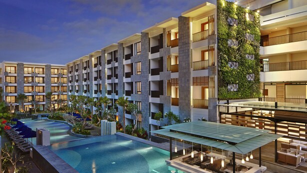 Un hotel moderno con una amplia piscina exterior, varios balcones y una terraza en la azotea.
