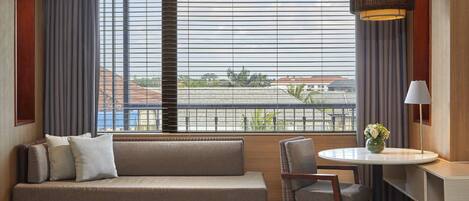 A modern hotel room with a sofa, a dining table with chairs, a lamp, and a view through large windows with blinds.