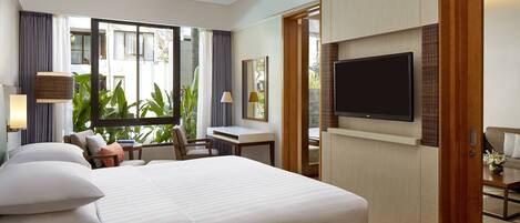 A hotel room with a large bed, a desk, a TV, and a view of greenery outside.