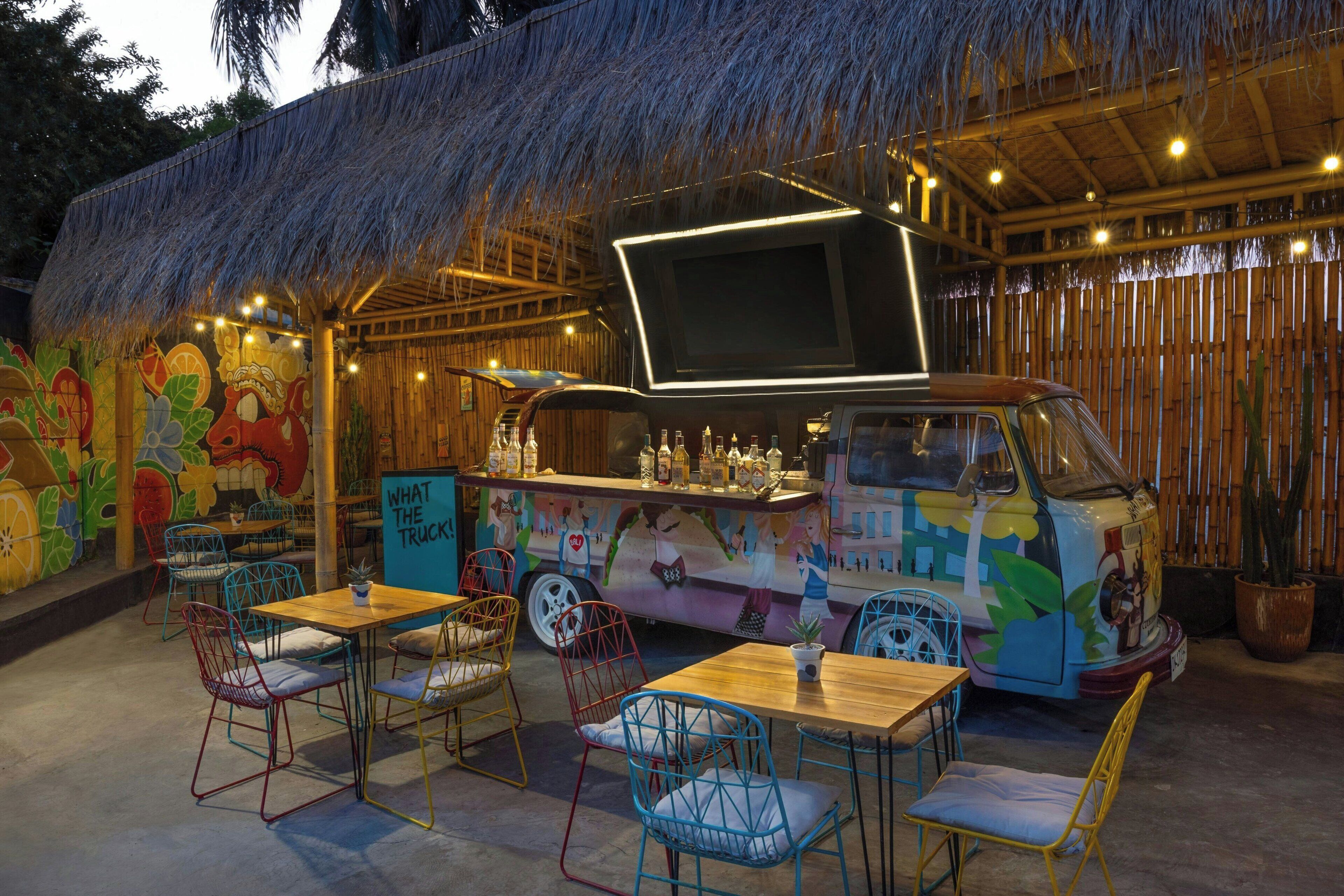 A food truck with a colorful mural, outdoor seating with metal chairs, and a thatched roof structure.