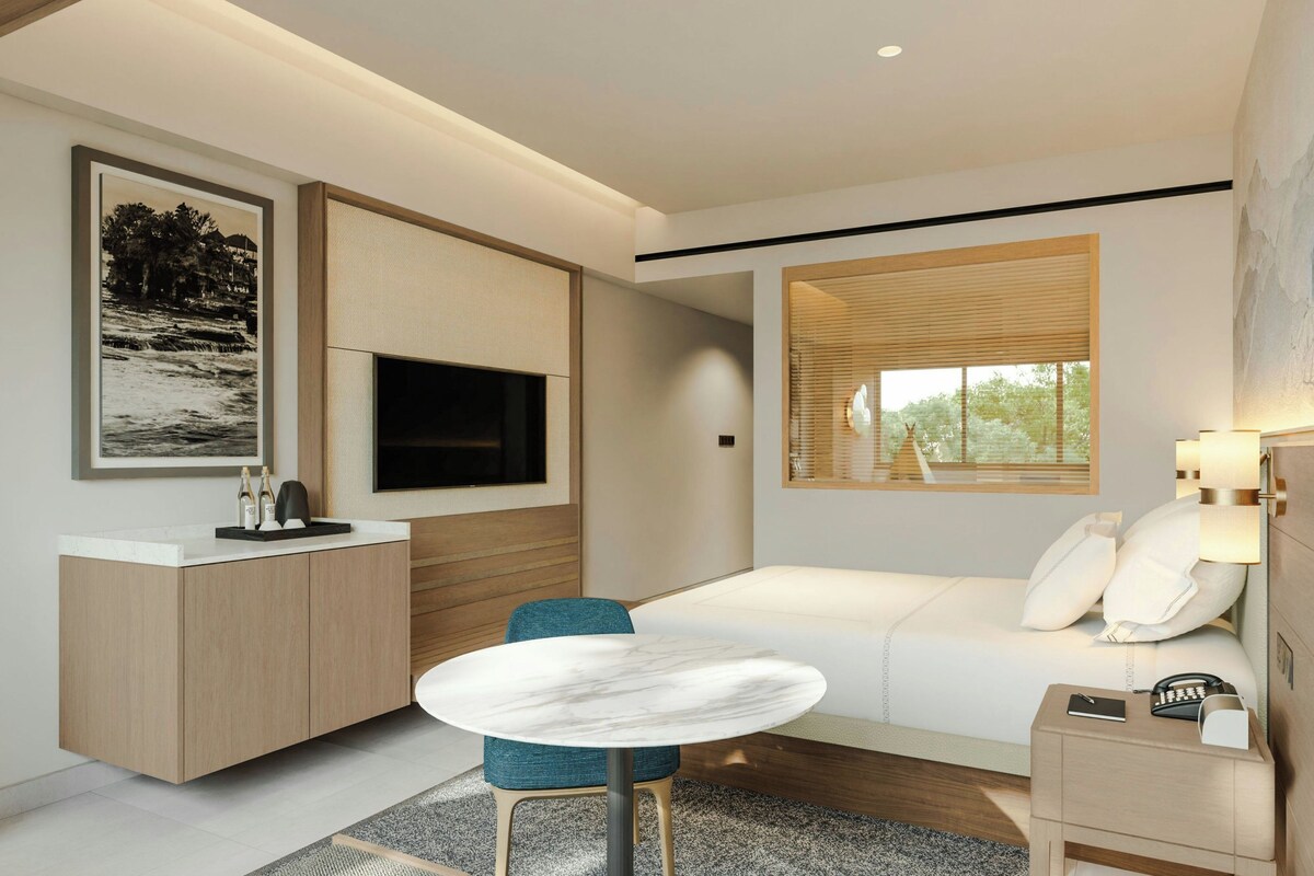 A modern hotel room with a bed, a desk, a TV, and a window with blinds.